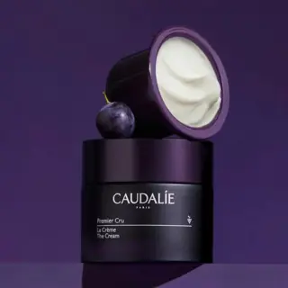 Caudalie Premier Cru cream jar with lid open, next to a grape, on a purple background.