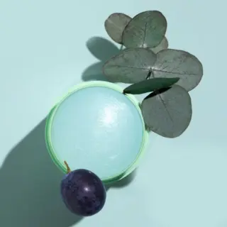Green jar with blue cream, eucalyptus leaves, and a purple plum on a light blue background.