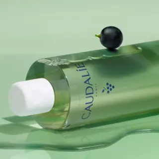 Green Caudalie bottle with white cap, grape and liquid on green surface.