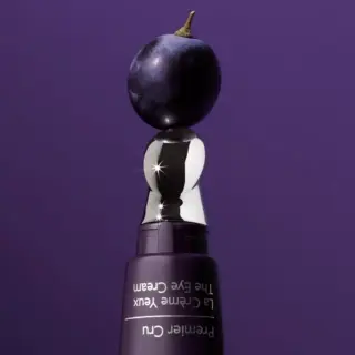 Purple eye cream bottle with a shiny silver cap, topped by a grape, on a purple background.
