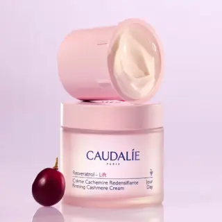 Caudalie Resveratrol Lift cream jar with lid off, grape beside, on a light purple background.