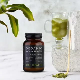 Kiki Health organic matcha powder jar beside a glass of matcha drink and green leaves.