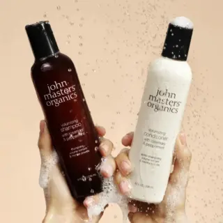 Two hands holding John Masters Organics shampoo and conditioner bottles with water droplets.