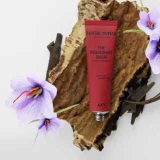 Red deodorant balm tube on bark with purple flowers. Text: Santal Nomad, AKT, saffron, sandalwood, leather.