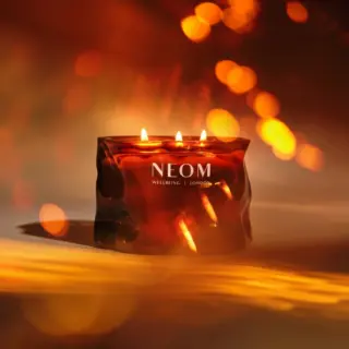 Lit candle in a dark amber glass with "NEOM Wellbeing London" text, warm glowing bokeh background.