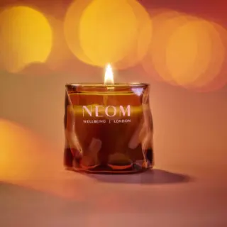 Lit NEOM candle with "Wellbeing London" text, set against a warm, blurred background.