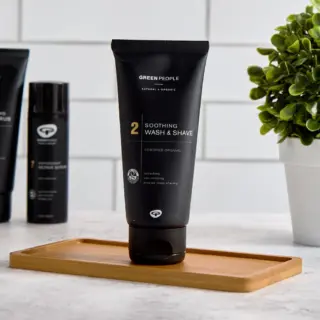Black tube of Green People Soothing Wash & Shave on a wooden tray, plant in background.