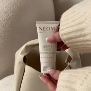 NEOM Energising Hand Balm, 30ml, held by a person in a cream jumper, near a beige bag.