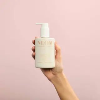 Hand holding NEOM lotion bottle against a pink background.
