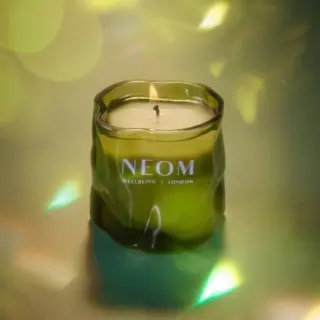 Green glass candle with "NEOM Wellbeing London" text, lit wick, on a reflective surface.