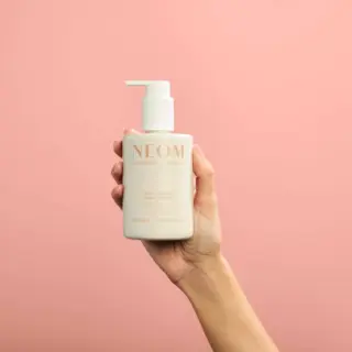 Hand holding NEOM hand lotion bottle against pink background.