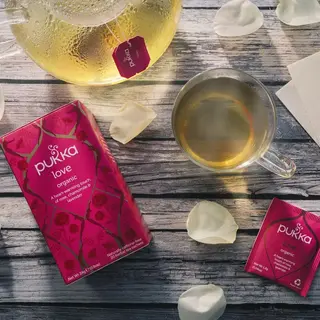 Pukka Love organic tea with teapot, cup, rose petals, and card on wooden surface.