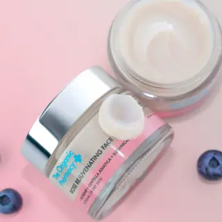 Two jars of rose rejuvenating face cream on a pink surface with blueberries.