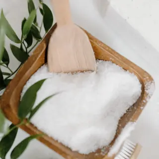 Wooden bowl of bath salts with a scoop, green leaves, and a bar of soap nearby.