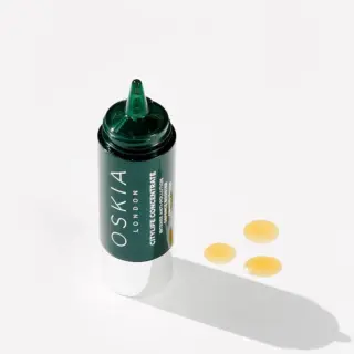 Oskia London skincare bottle with three yellow serum drops on white background.