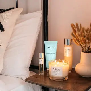 Neom products on bedside table with lit candle and dried flowers.