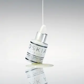 Oskia Isotonic Hydra-Serum bottle with liquid pouring, reflected on a glossy surface.