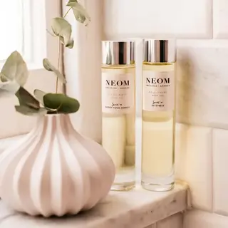 Two Neom Organics body oils on a marble shelf beside a white vase with green leaves.