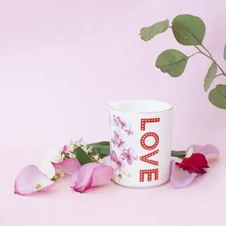 White jug with "LOVE" text, floral design, surrounded by pink petals on a pink background.