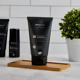 Green People Itch Away Shampoo tube on a wooden tray, with a plant and other products nearby.