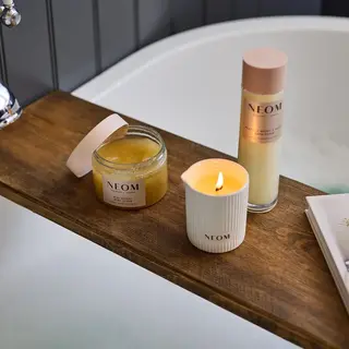 Neom body scrub, bath foam, and lit candle on wooden bath tray.