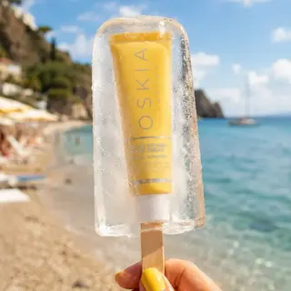 Hand holding ice lolly with Oskia skincare tube inside, beach and sea in background.