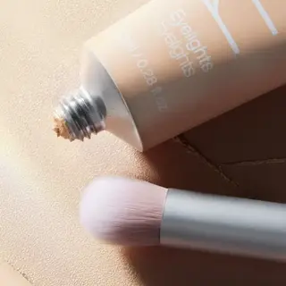 Open tube of Eyelights cream with a makeup brush on a textured surface.