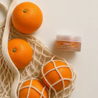 Oranges in a net bag next to a jar of SUPER-C beauty capsules on a light background.