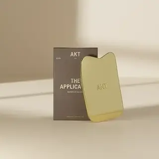 Gold AKT applicator with packaging on a beige surface.