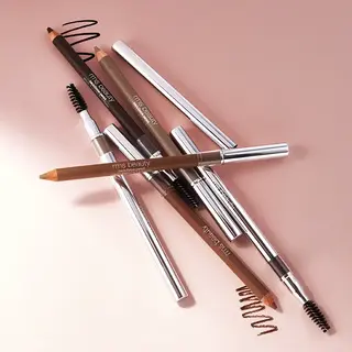 Assorted RMS Beauty brow pencils on a pink surface.