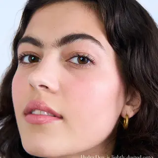 Woman with glowing skin, wearing gold hoop earring. Text: "Hydra Dew is lightly dusted...