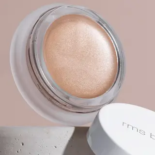 Open jar of shimmering peach cream with "rms beauty" on the white lid, set on a stone surface.
