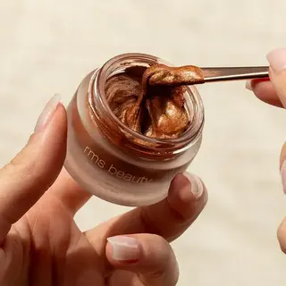 Hands holding a jar of rms beauty bronze cream with a spatula.