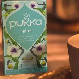 Pukka Relax herbal tea box with chamomile, fennel, and marshmallow root, next to a cup on a woven mat.