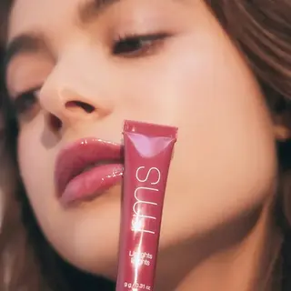 Woman holding a pink lip gloss tube labelled "SUU" near her face.