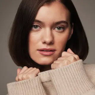 Woman with short brown hair in a beige jumper, holding collar near face. Text: "TAN".