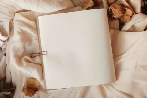 Open blank notebook on beige fabric with dried leaves.