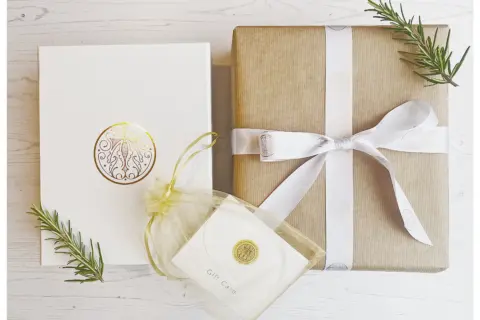Two gift boxes with rosemary sprigs and a gift card in a sheer pouch on a white wooden surface.