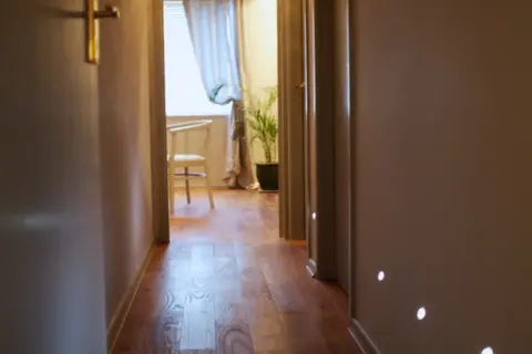 Hallway with wooden floor, open door, and a lit room with a chair and plant visible at the end.
