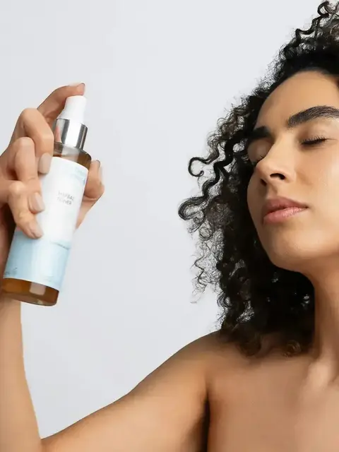 Person holding a bottle of herbal toner, eyes closed, curly hair.