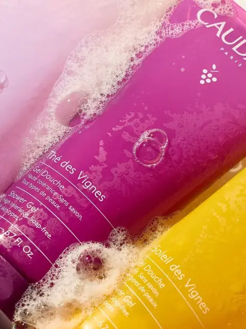 Three Caudalie shower gels in purple, pink, and yellow, covered in bubbles.