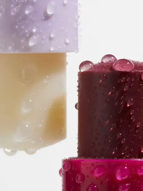Two lipsticks with water droplets, one beige and one dark red, on a white background.