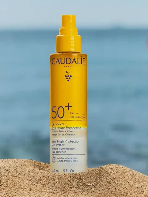 Caudalie SPF 50+ sun spray on sand with sea background.