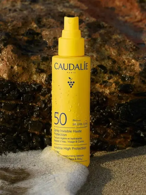Yellow Caudalie SPF 50 spray bottle on a sandy beach with rocks in the background.
