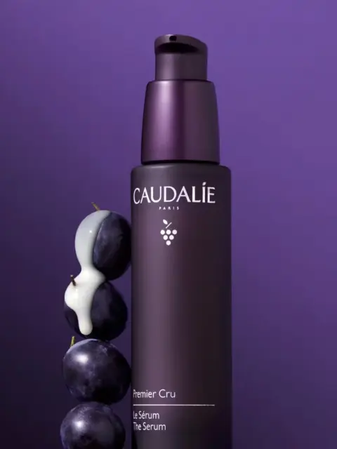 Caudalie Premier Cru Serum bottle with grapes and cream on purple background.