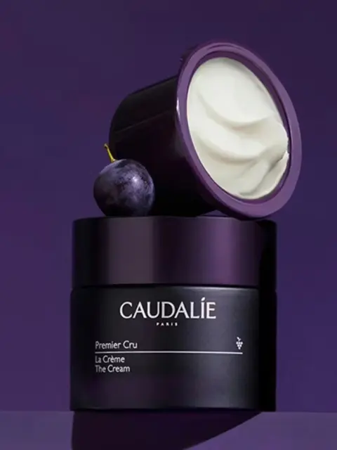 Caudalie Premier Cru cream jar with lid open, next to a grape, on a purple background.