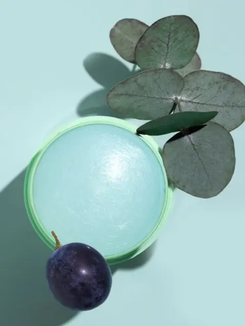 Green jar with blue cream, eucalyptus leaves, and a purple plum on a light blue background.