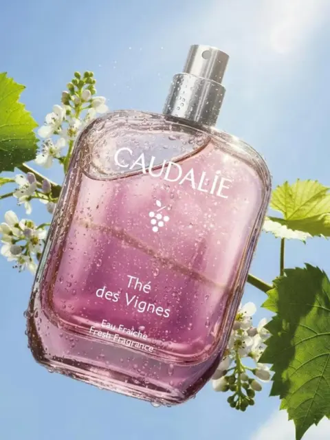 Caudalie Thé des Vignes fragrance bottle with water droplets, surrounded by green leaves and flowers.