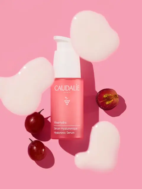 Pink Caudalie serum bottle with grapes and cream blobs on pink background.
