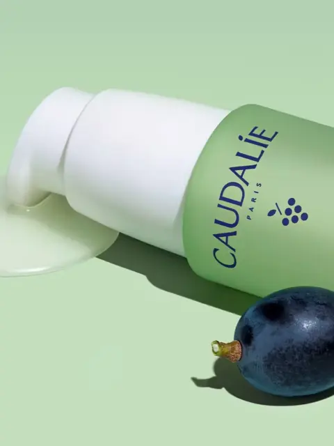 Caudalie Vinopure bottle with pump, liquid spill, and a grape on a green background.
